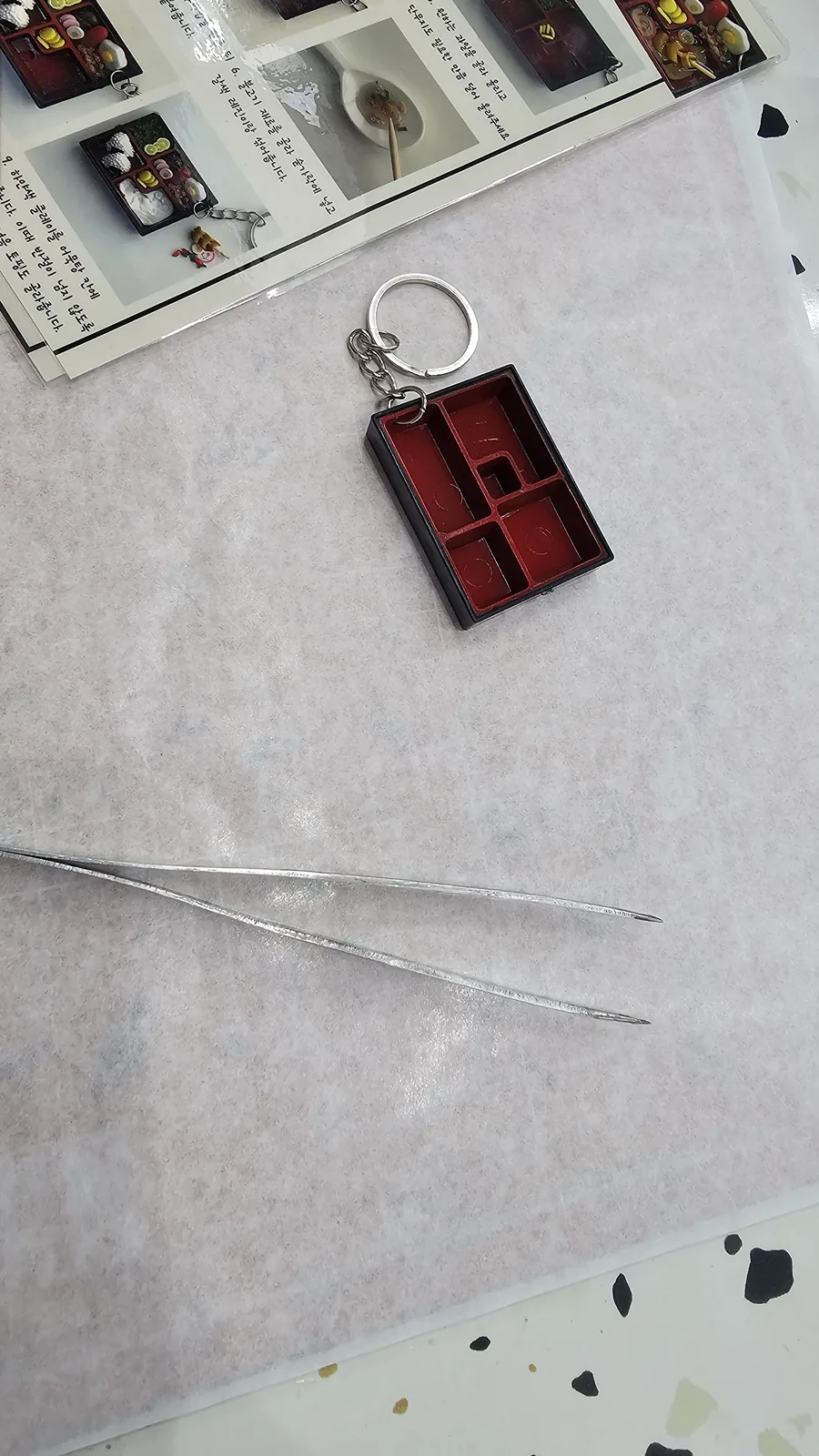Empty lunchbox keyring acrylic frame and tweezers on a white table, before the Amado class begins