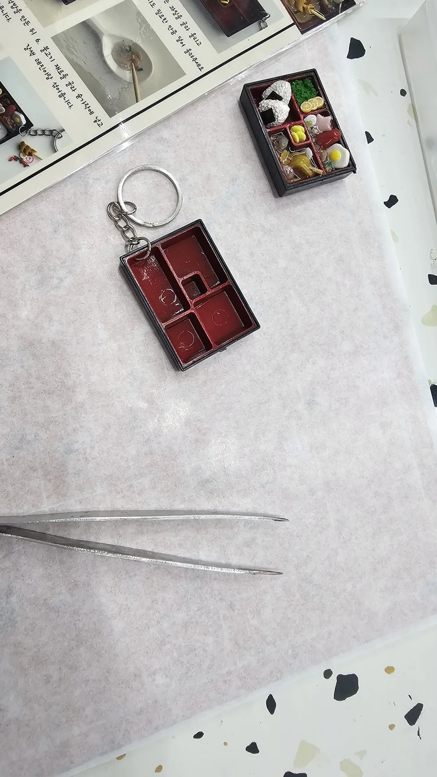 Empty lunchbox keyring frame beside a completed sample keyring with miniature rice balls, tweezers on the side