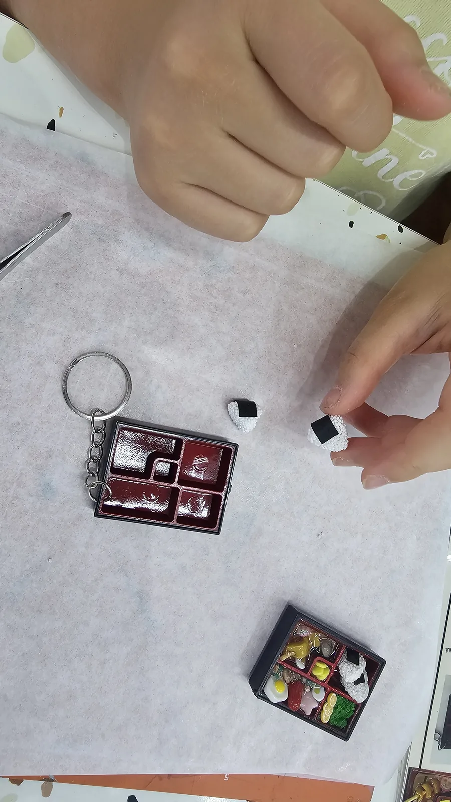 Hands placing a white clay rice ball into the lunchbox keyring frame, with a completed sample nearby