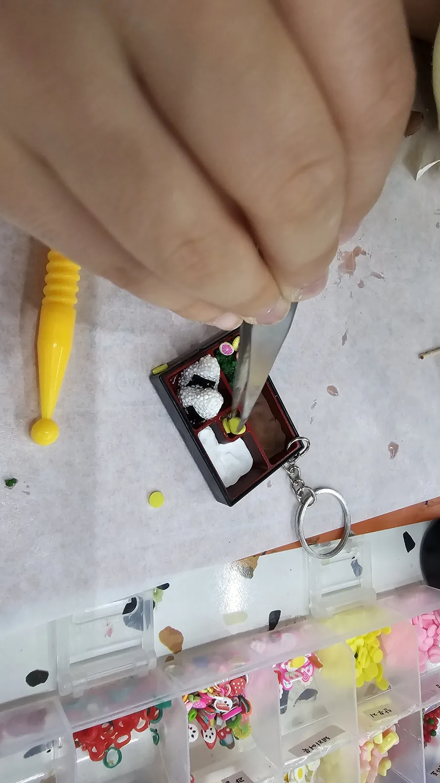Tweezers carefully placing miniature food pieces into the lunchbox keyring, miniature ingredient boxes below
