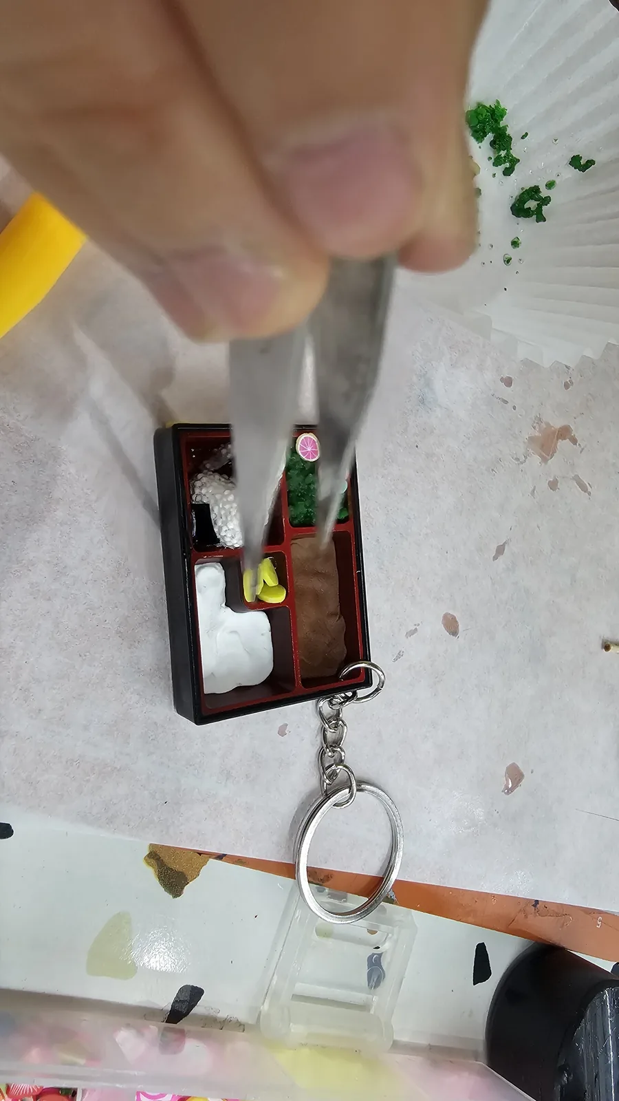 Close-up of lunchbox keyring with tweezers placing pink fish cake (eomuk) slices, rice ball and broccoli already in place