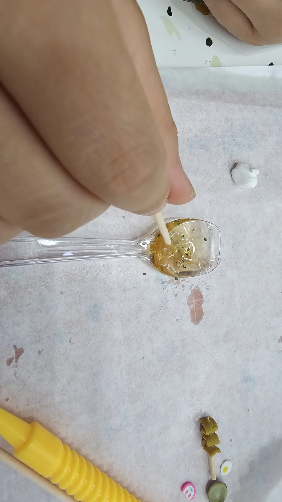 Hands stirring golden UV resin in a clear spoon to make miniature sauce for the lunchbox keyring