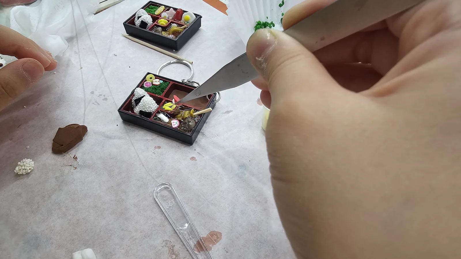 Tweezers adding small brown bulgogi meat pieces into the lunchbox keyring, completed sample keyring beside it
