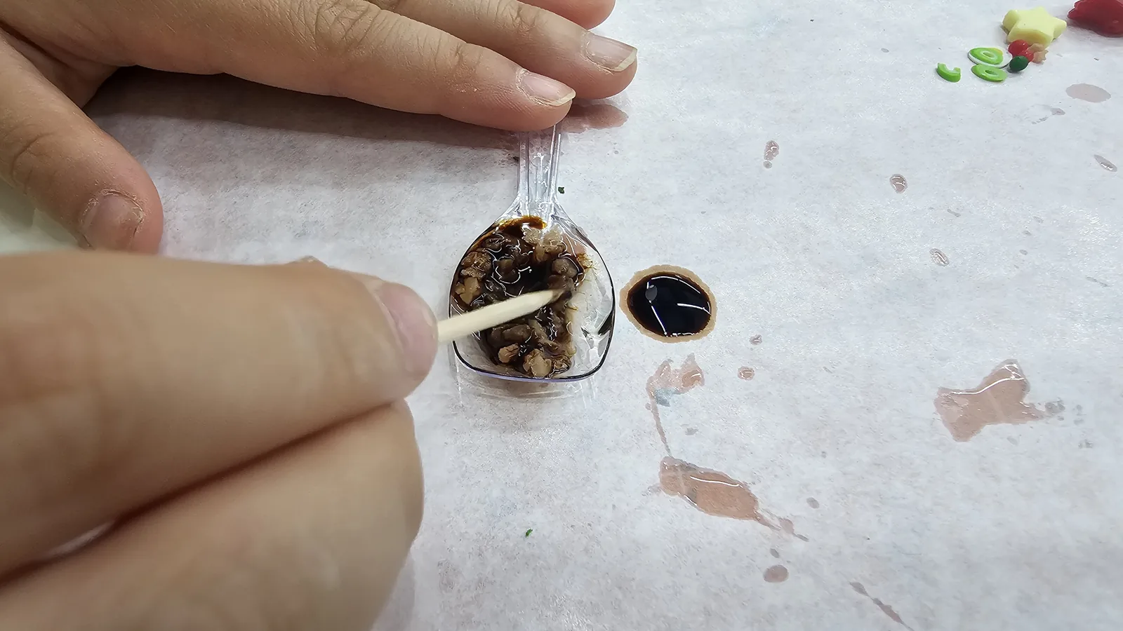 Mixing brown UV resin with small ingredient pieces in a clear spoon to create miniature bulgogi sauce