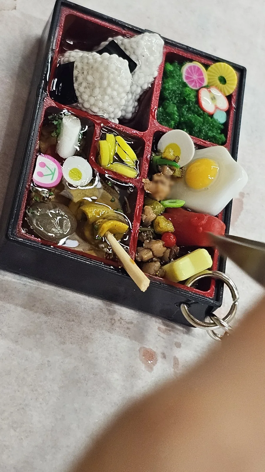Nearly completed lunchbox keyring close-up with tweezers placing the last food pieces, rice ball and bulgogi filling visible