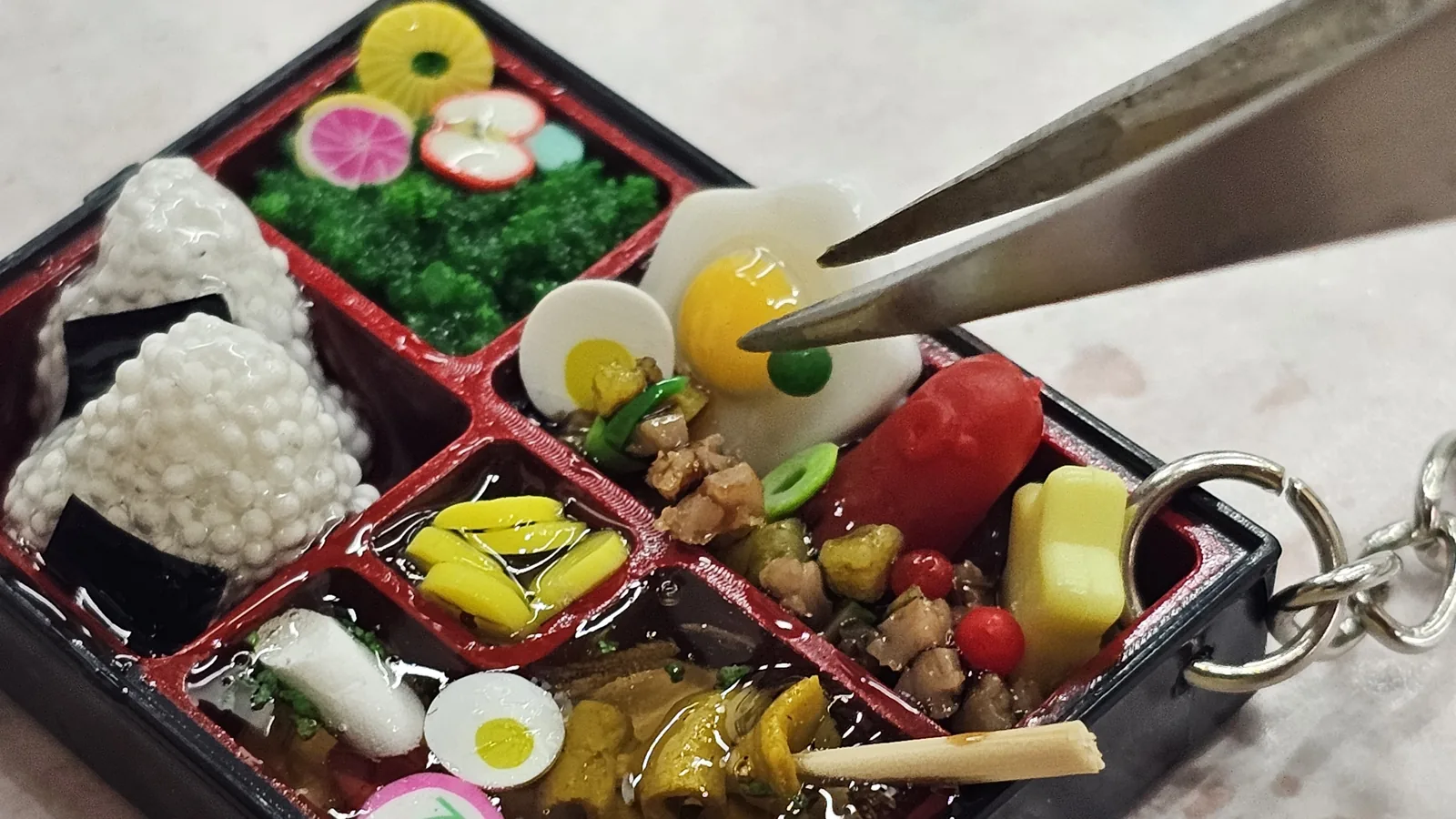 Close-up of lunchbox keyring with tweezers placing a tiny fried egg, the box nearly full with colorful miniature food