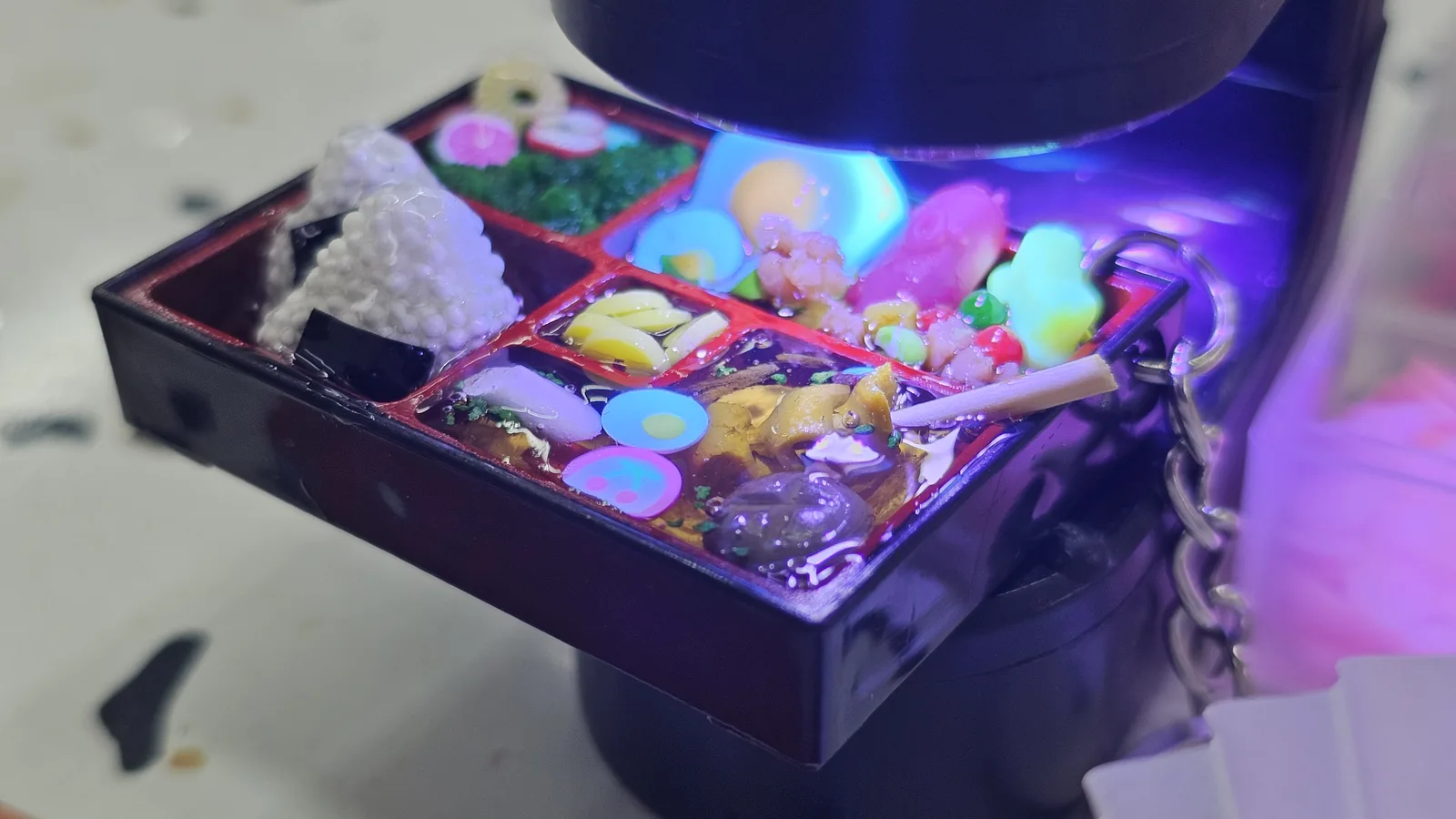 Lunchbox keyring glowing under purple-blue UV lamp light during the resin curing step at Amado
