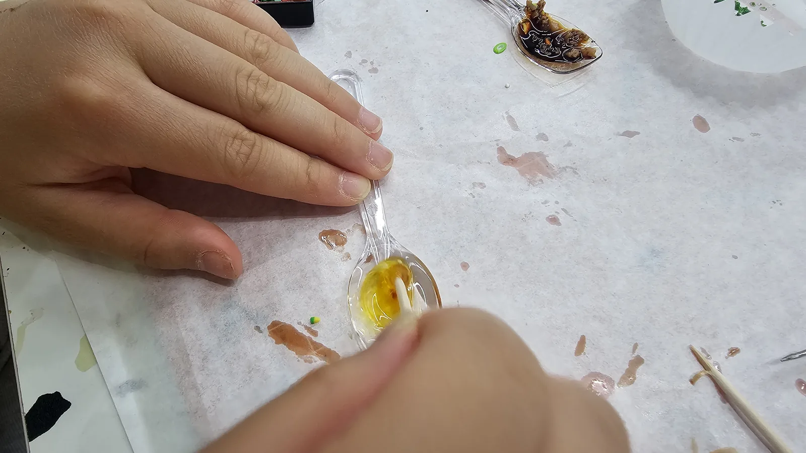 Two hands stirring golden UV resin in a clear spoon, creating miniature sauce for the lunchbox keyring