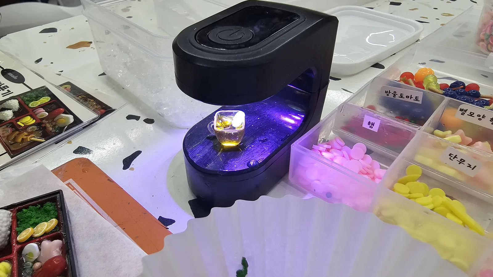UV lamp curing a small miniature resin cup, ingredient boxes for tomato, ham, and radish visible nearby
