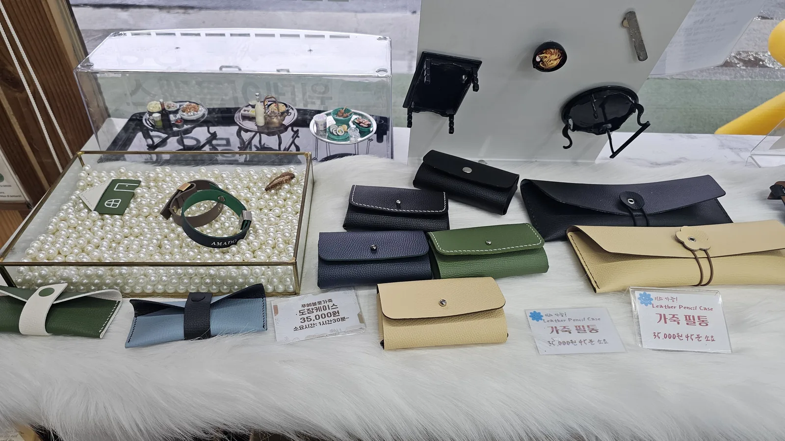 Display of leather stamp cases, pencil cases, and AMADO-engraved leather bracelets beside a piano-shaped glass display cabinet
