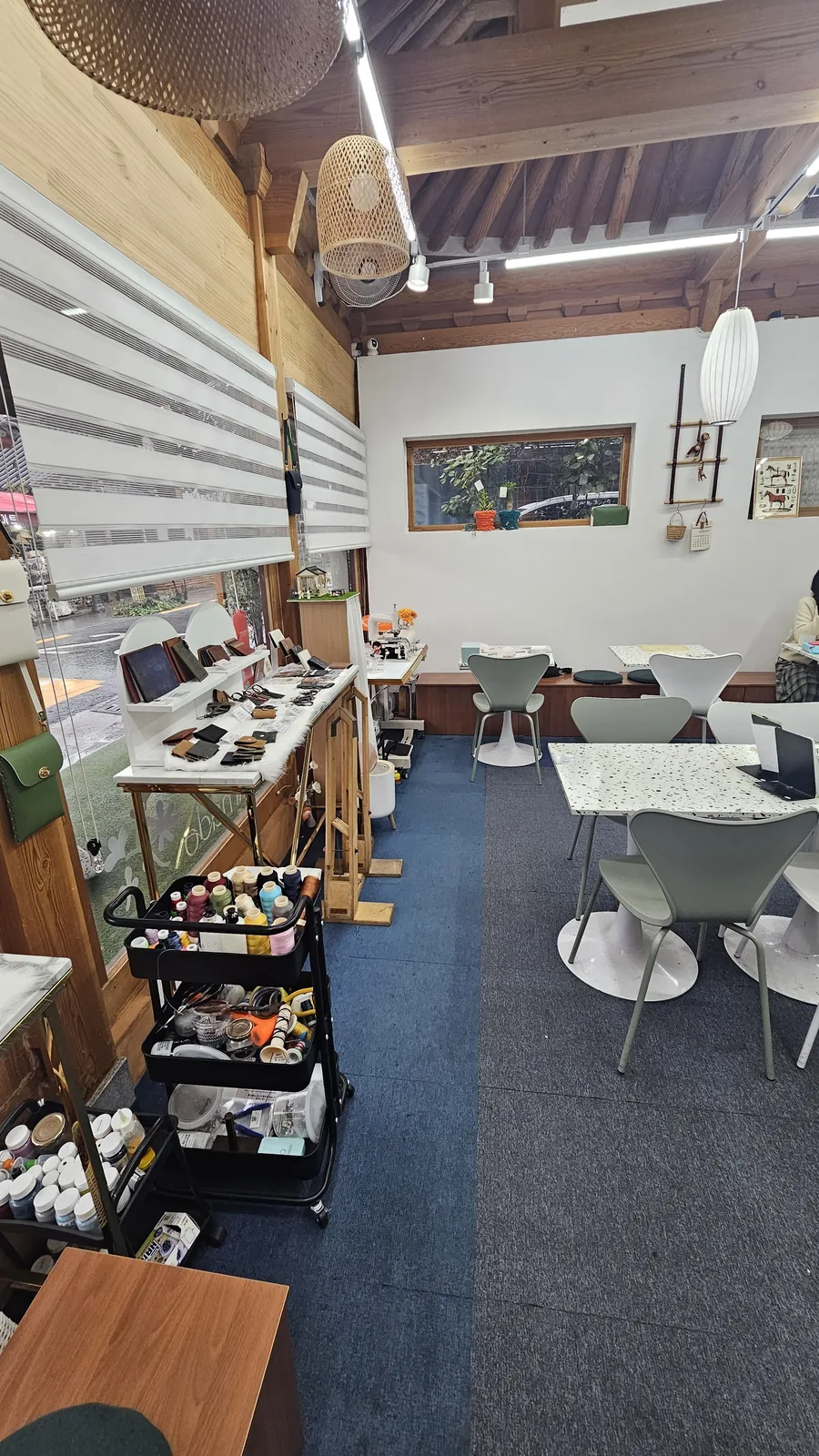Wide interior view of Amado Leather Workshop with hanok ceiling, leather worktables, and material cart, person partially visible at right