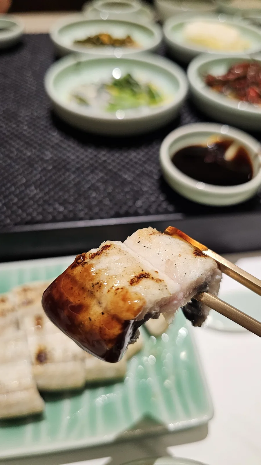 Close-up of grilled eel piece held with chopsticks at Daedong Jangeo with banchan in background