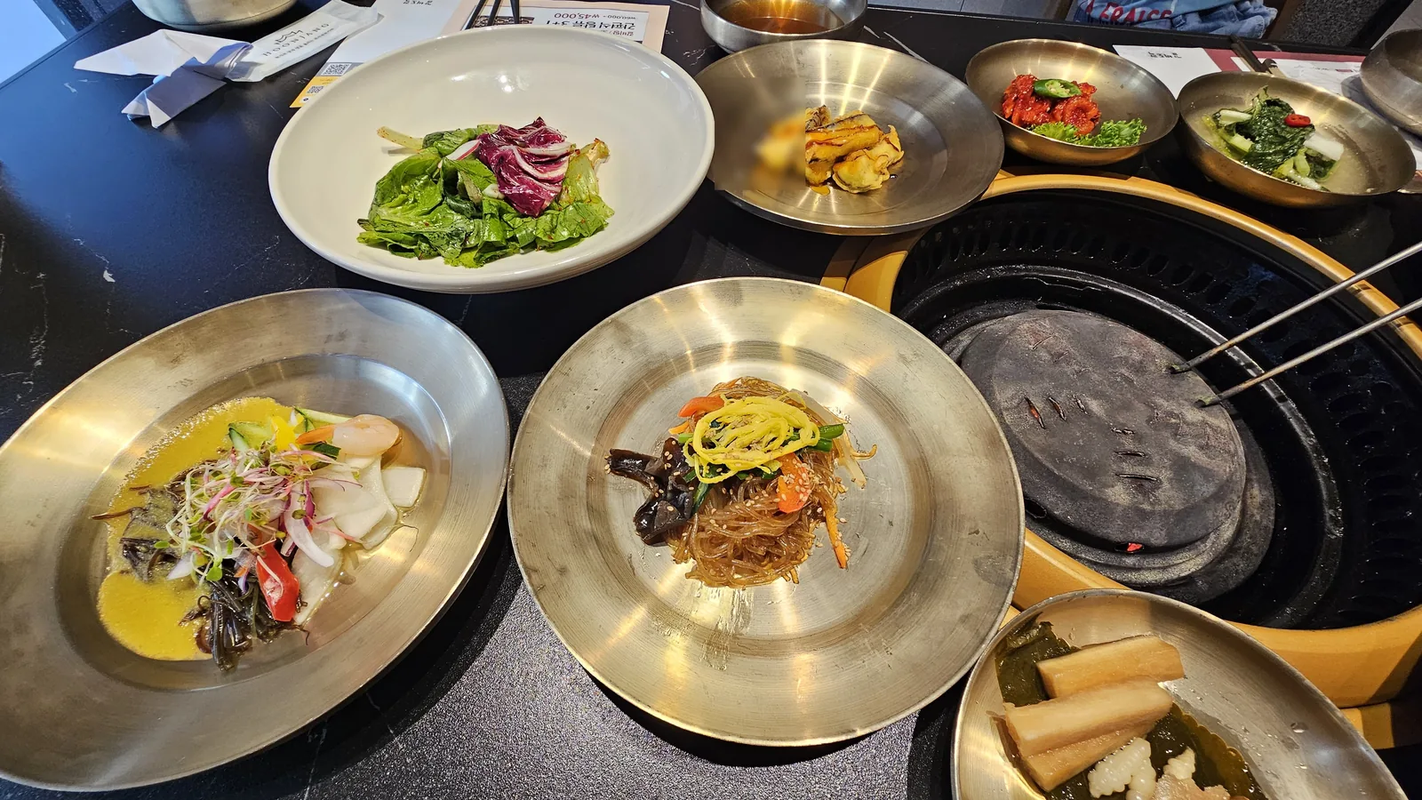 Banchan spread at Galbidorak — japchae, seasoned greens, kkakdugi, pumpkin salad and charcoal grill table setup