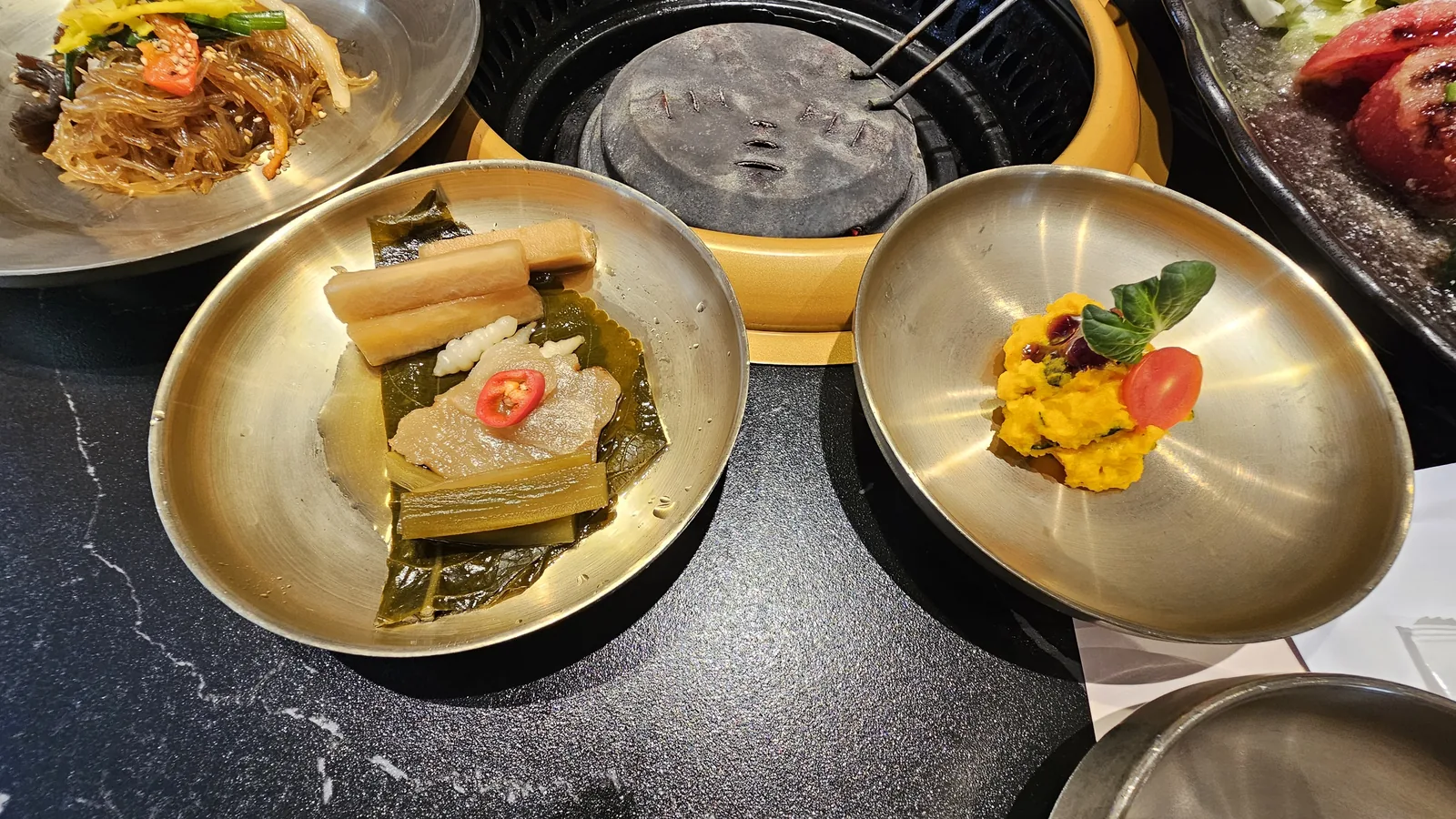 Galbidorak banchan close-up — stainless steel bowls with dried kelp, radish, tofu kimchi and candied pumpkin