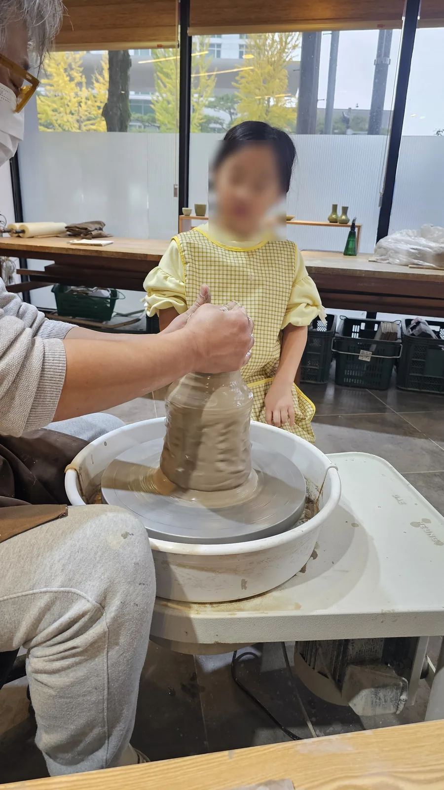 Young girl sitting at pottery wheel getting started with instructor's hands-on guidance at Udoye Gongbang