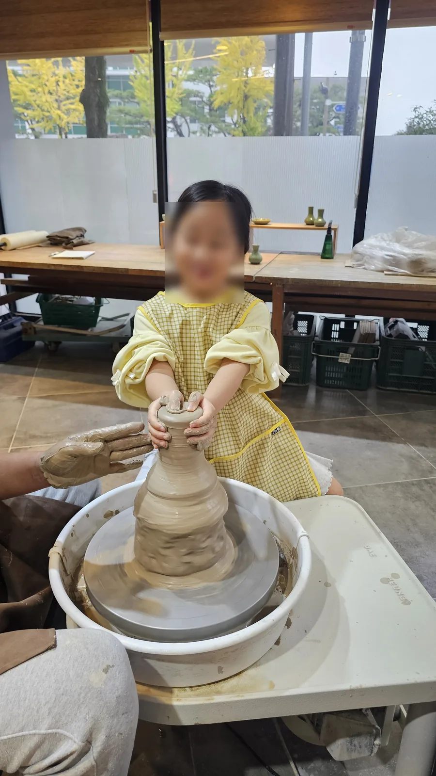 Young girl smiling while shaping clay on the pottery wheel with instructor assistance at Udoye Gongbang