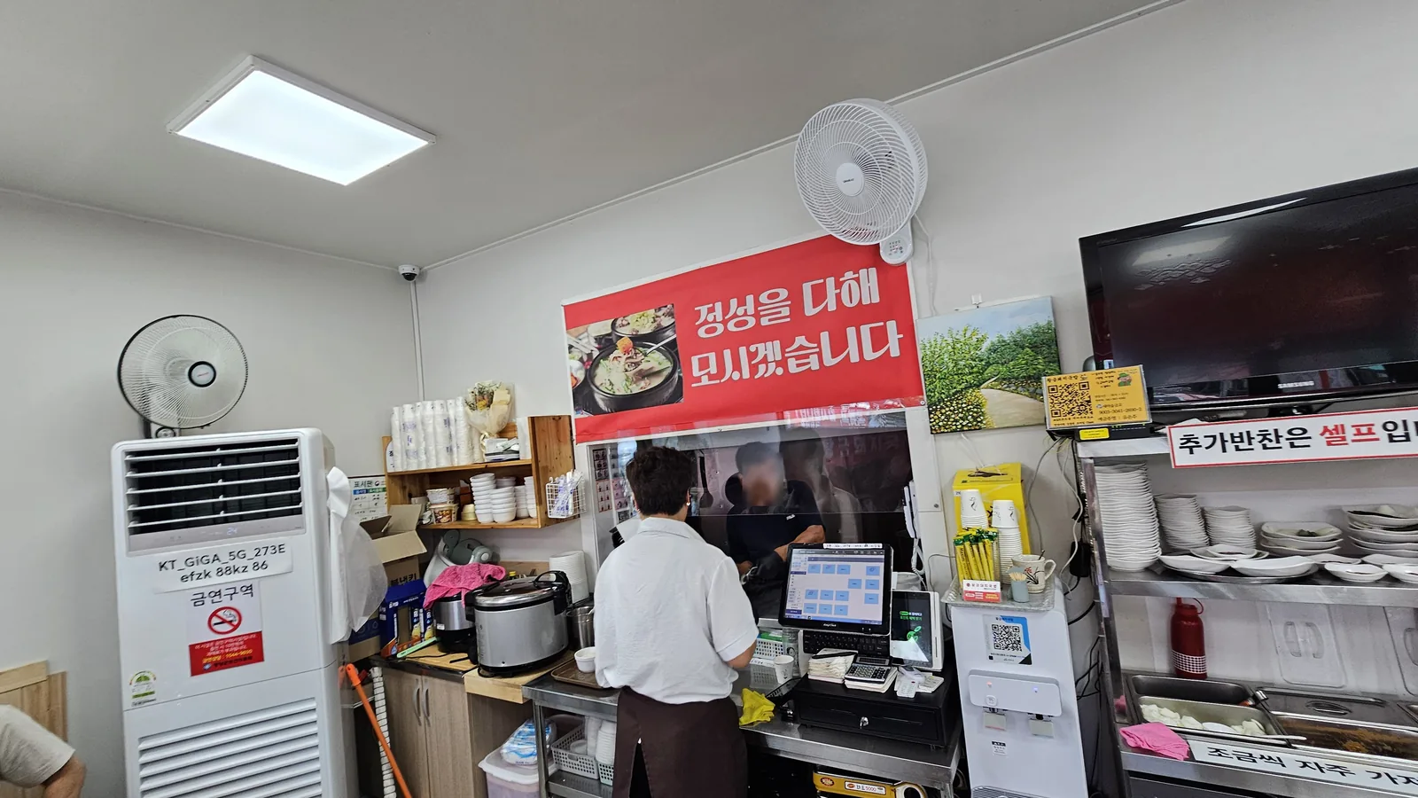 Kitchen counter and staff at Hwanggeum Dwaeji Gukbap with self-service side dish station signage