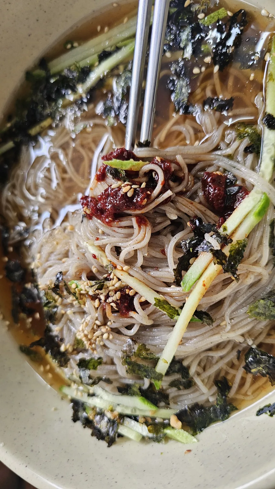 Chopsticks lifting fresh buckwheat noodles from a makguksu bowl with gochujang chili paste on the side
