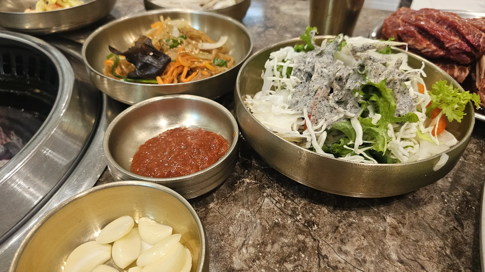 Top-down view of Jogane Galbi table — japchae, black pepper dressed salad, ssamjang, garlic, and raw galbi visible