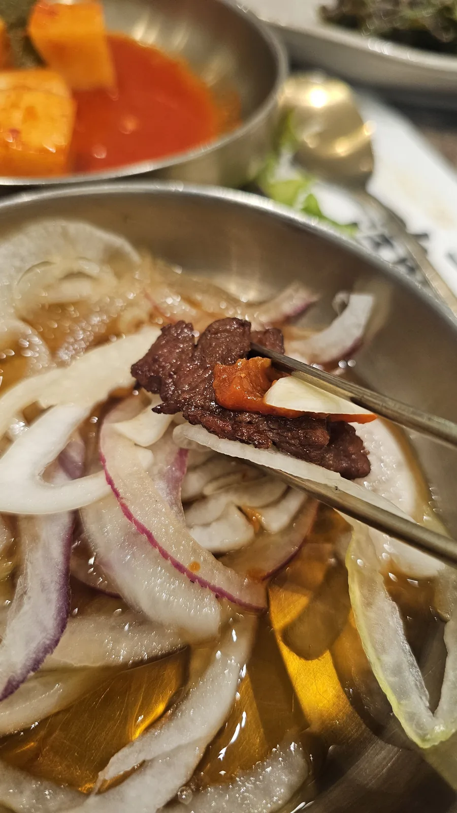 Chopsticks picking up a grilled galbi piece over a bowl of pickled onions at Jogane Galbi Mokdong