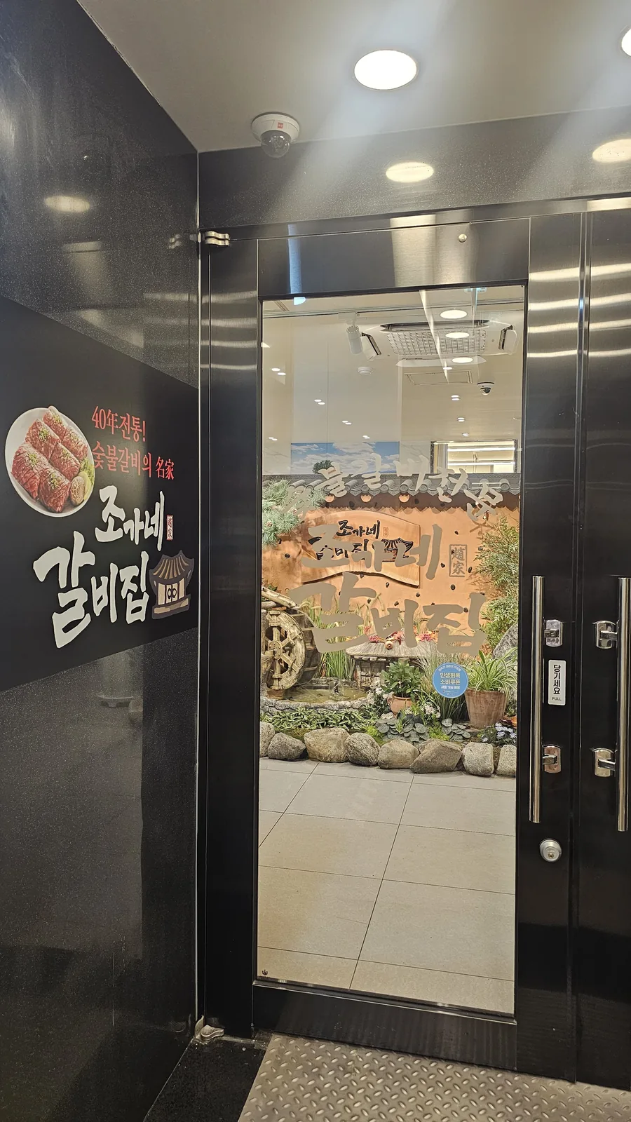 Exterior entrance of Jogane Galbi Mokdong with restaurant signage and view through glass doors into indoor garden