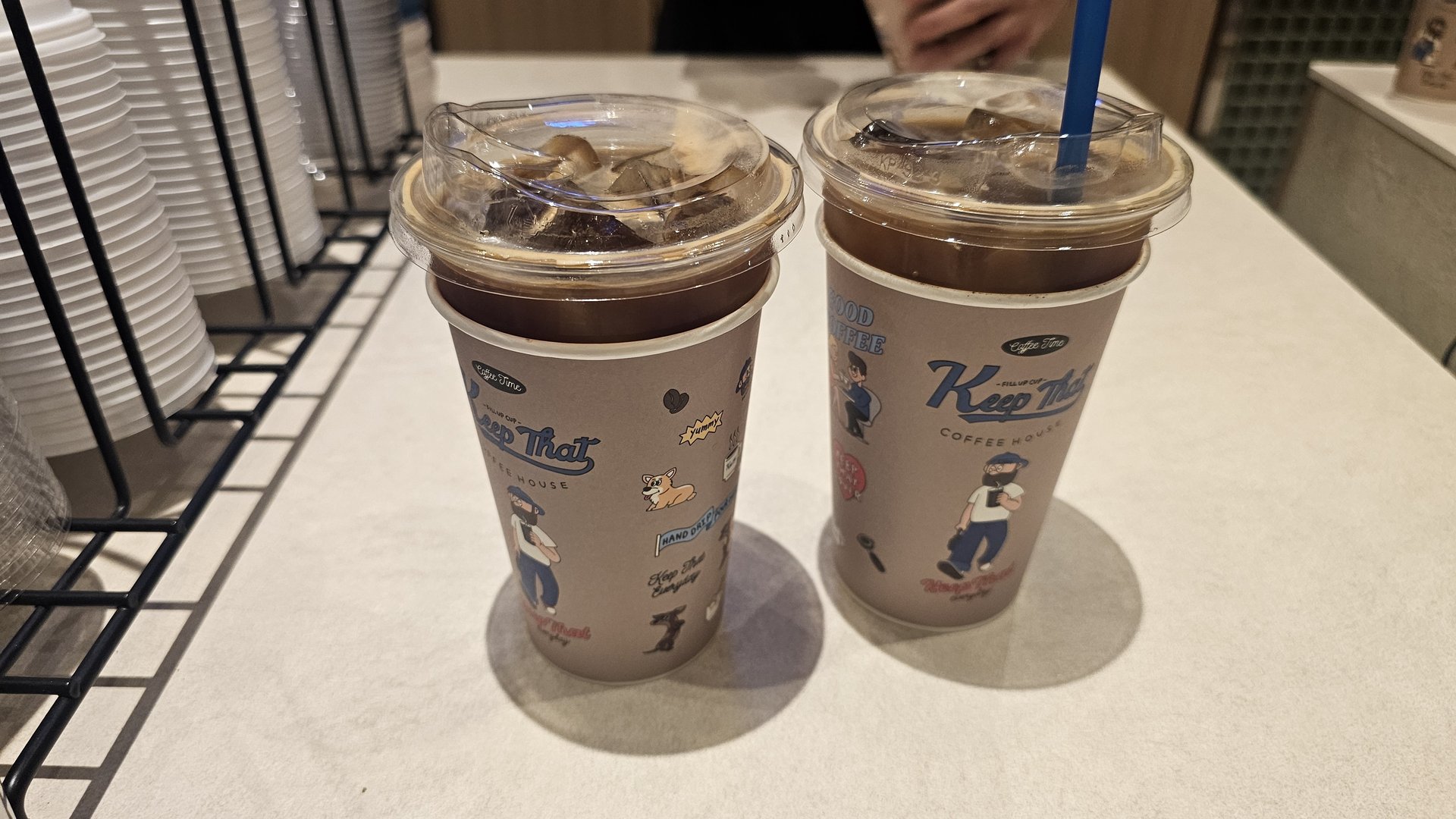 Two iced Americanos in Keep That Coffee House logo paper cups lined up on the counter