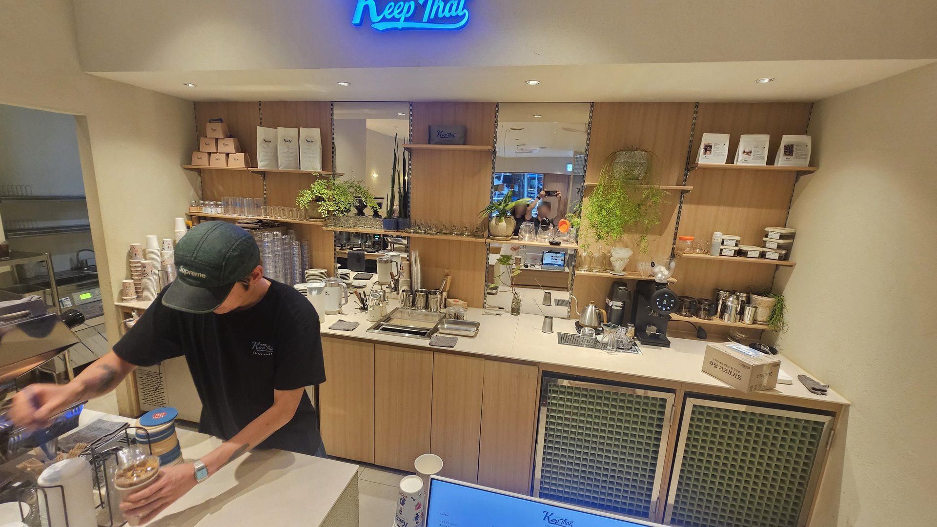 Barista preparing drinks at the bar counter with wood-tone interior and neon Keep That logo sign
