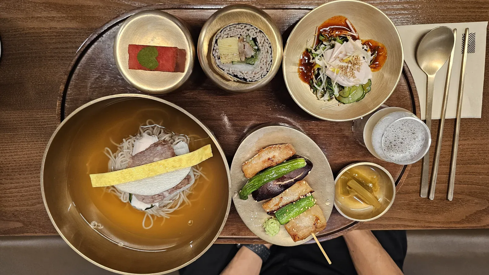 Meomil Danpyeon bansang tray with cold buckwheat noodles in clear broth, chicken skewer, buckwheat kimbap, and sikhye drink
