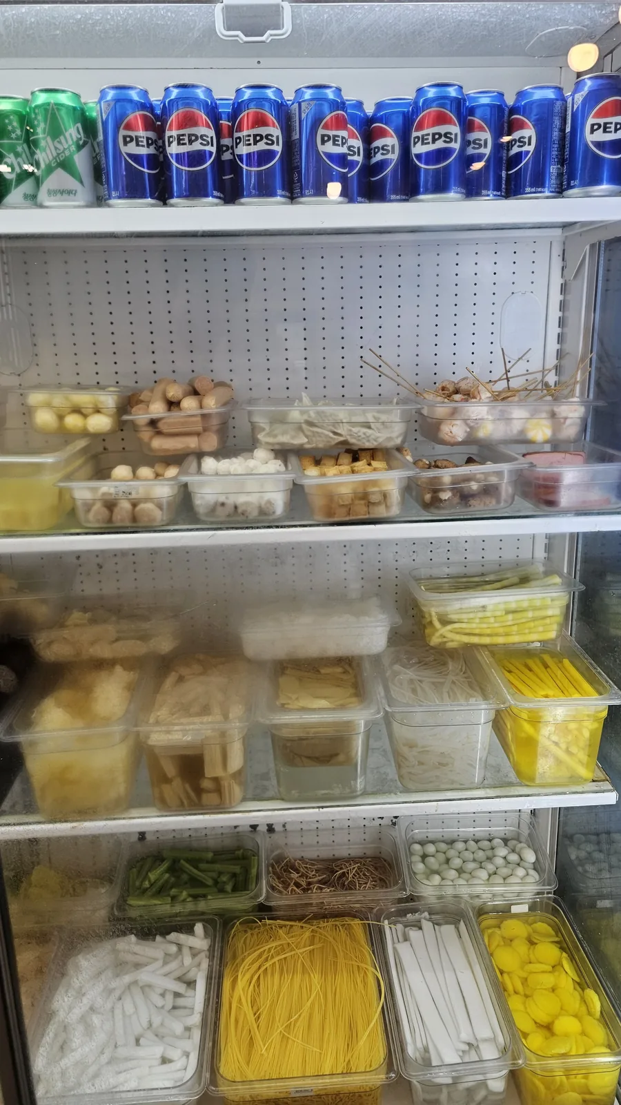 Malatang ingredient fridge showing fish cake balls, noodles, rice cakes, quail eggs, and assorted vegetables