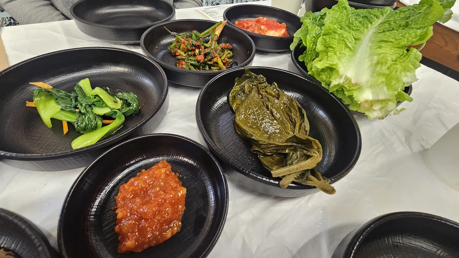 Korean banchan side dishes including seasoned bok choy, red pepper seasoning, pickled perilla leaves, cabbage wrap leaves, and red sauce