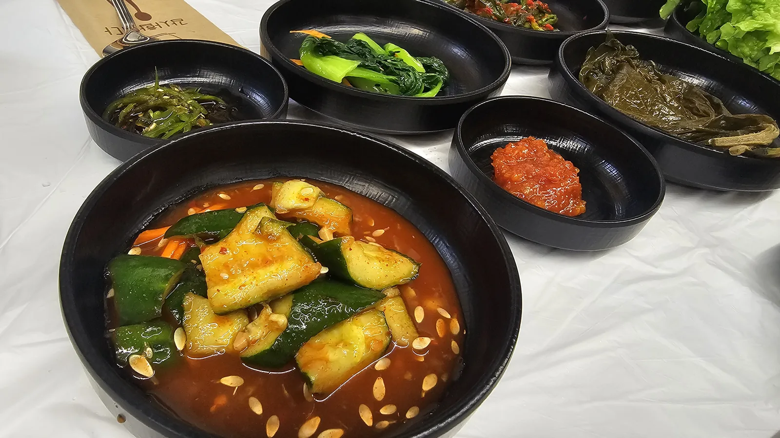 Full spread of Korean banchan side dishes including braised cucumber at front, seaweed, bok choy greens, pickled perilla, and lettuce wraps