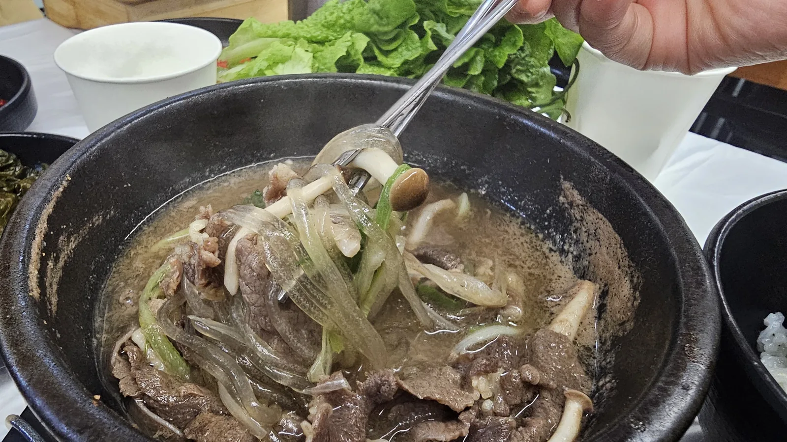 Mid-meal shot of ttukbaegi stone pot galbi soup with chopsticks pulling glass noodles and rib meat