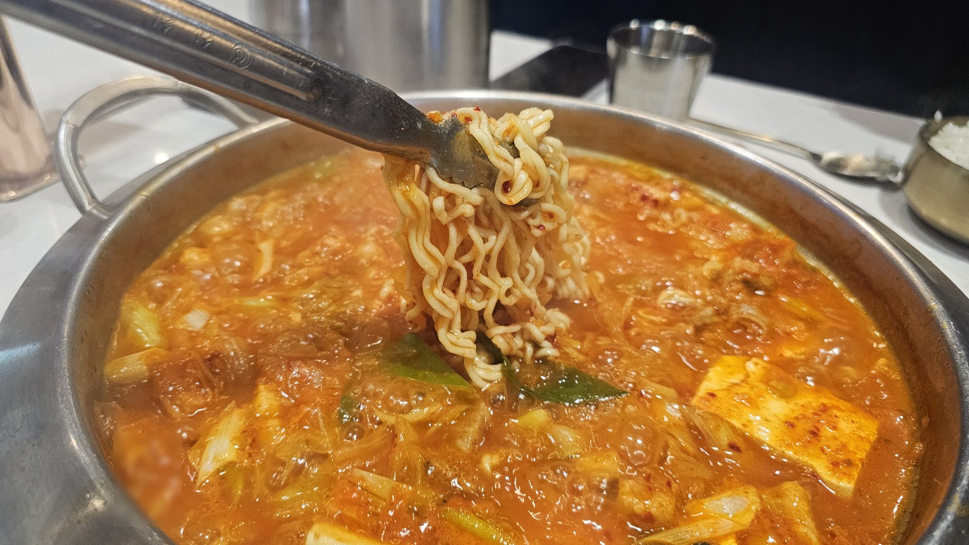 Close-up of lifting ramen noodles from kimchi jjigae with tongs showing rich red broth at Shindosegi