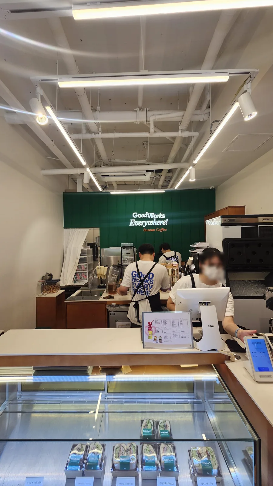 Order counter at Sunsun Coffee Yeouido with staff preparing drinks, sandwich packages in glass display below, GoodWorks Everywhere Sunsun Coffee wall text